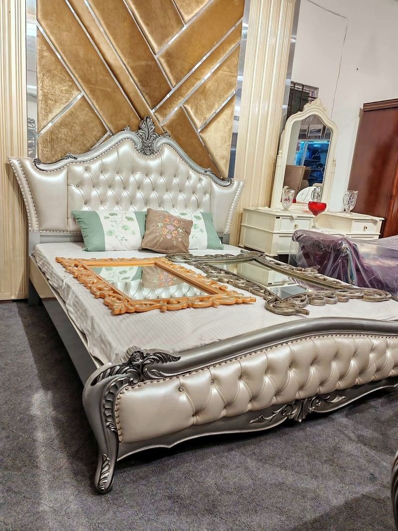 Pearl-coloured bed with a tall patterned cushioned headboard and carved frame